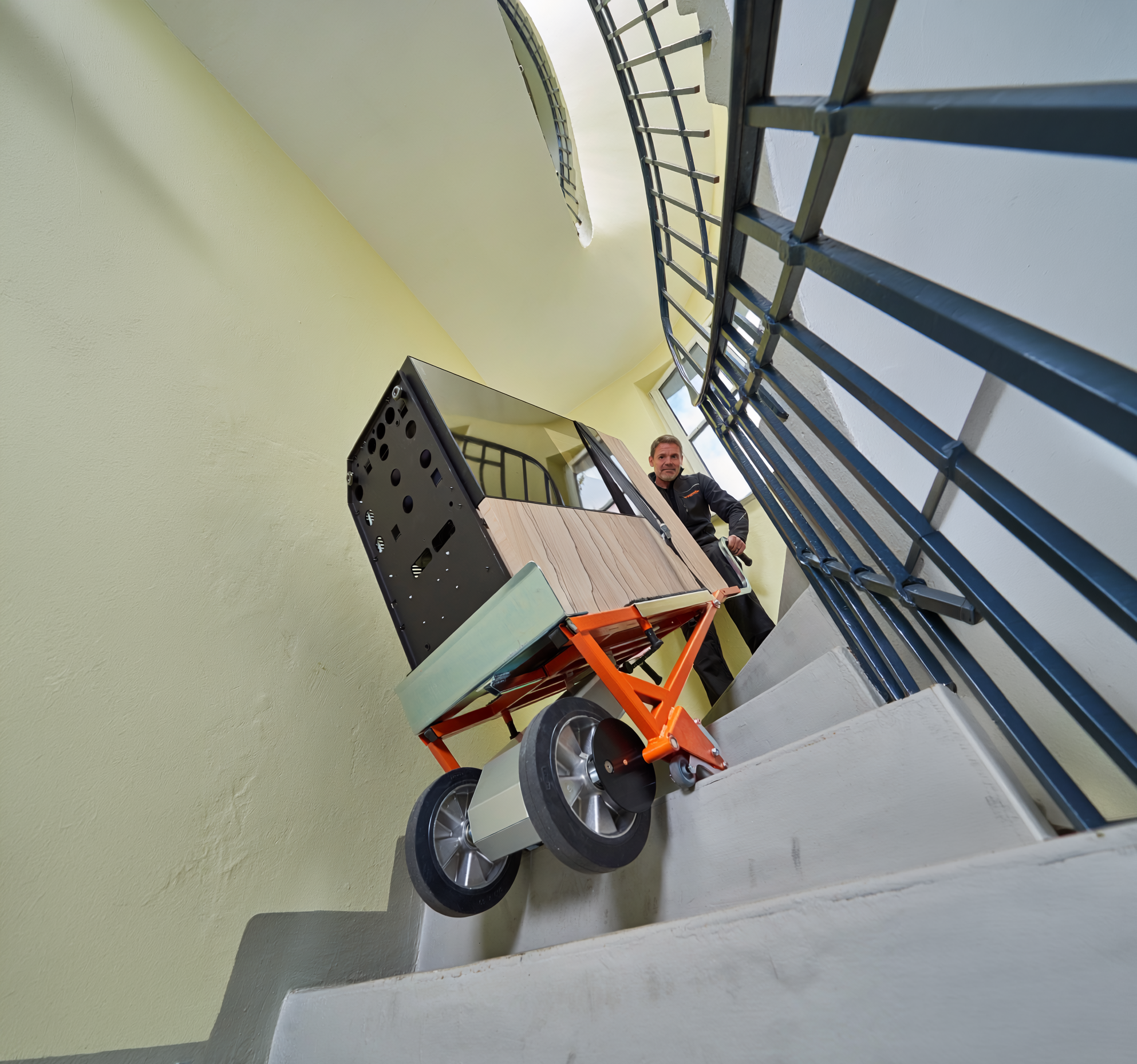 Das Bild zeigt einen Mann, der mit Hilfe eines CargoMaster Treppensteigers für Lasten einen Ofen über eine Wendeltreppe transportiert. Zu sehen sind außerdem die Steigräder des Treppensteigers sowie die Sicherheitsbremsen, die an der Treppenkante automatisch gestoppt haben.
