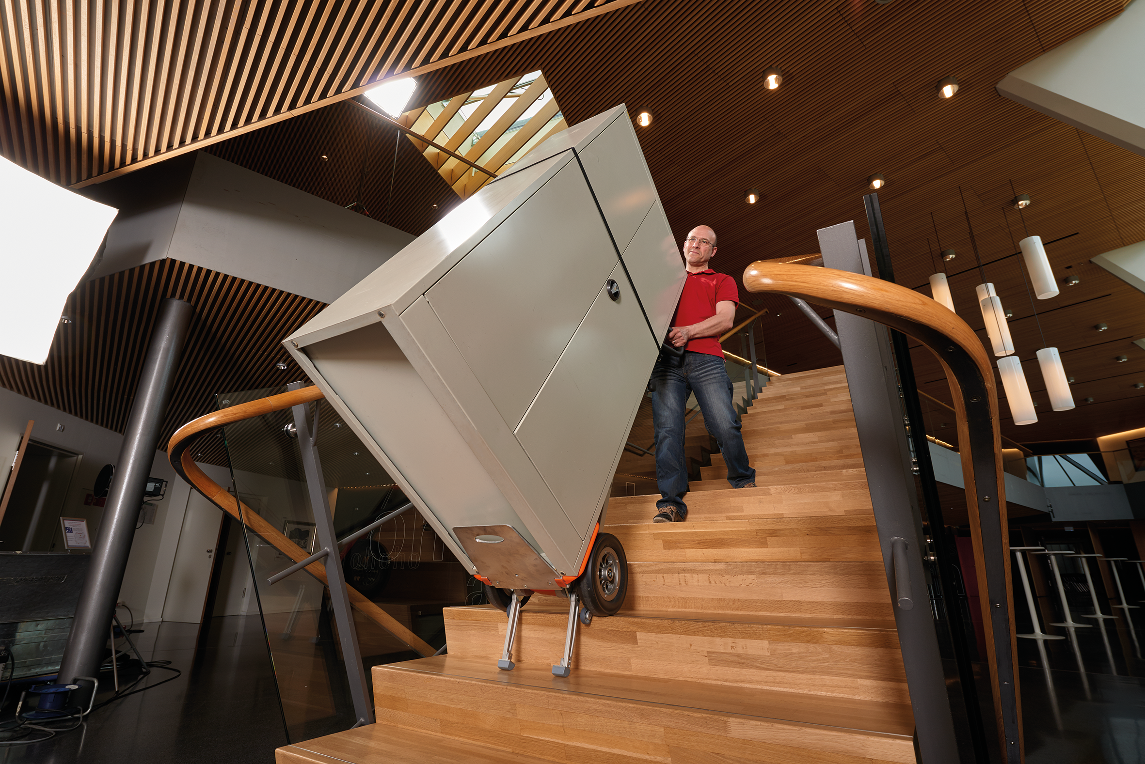 Das Bild zeigt einen Mann mittleres Alters, der mit Hilfe eines CargoMaster Treppensteiger für Lasten einen großen grauen Schrank auf einmal über Treppen befördert. Der Mann sieht dabei sehr entspannt aus und braucht kaum Kraft.